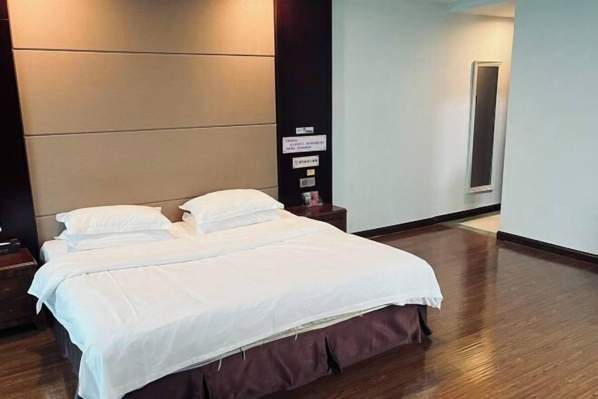 A hotel room with a large bed, a nightstand, a wall-mounted control panel, and a door.