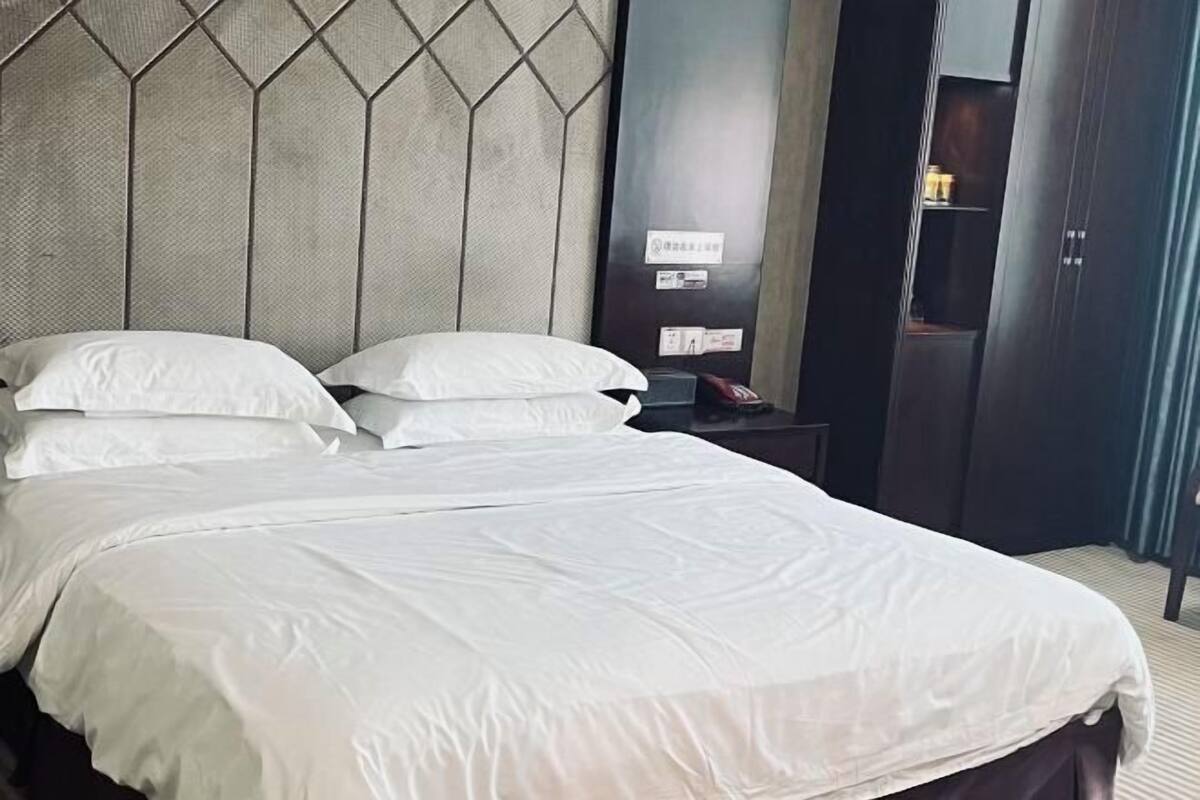 A hotel room with a large bed, a headboard with a geometric pattern, a nightstand with a telephone, and a wardrobe.