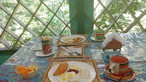 Ein Tisch ist reichhaltig mit Speisen gedeckt, darunter Eier, Brot und Obst, in einem Raum mit einem Gitterfenster und einer blumigen Tischdecke.