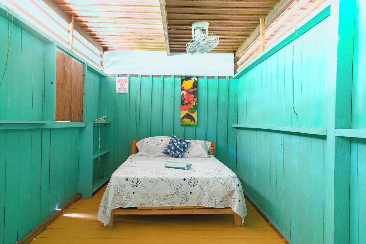 Une petite pièce étroite aux murs de couleur turquoise et au plafond en bois. Elle contient un lit simple recouvert d’une courtepointe blanche à motifs floraux et agrémenté d’un coussin coloré. Un ventilateur est fixé au plafond, et un tableau coloré est accroché au mur.