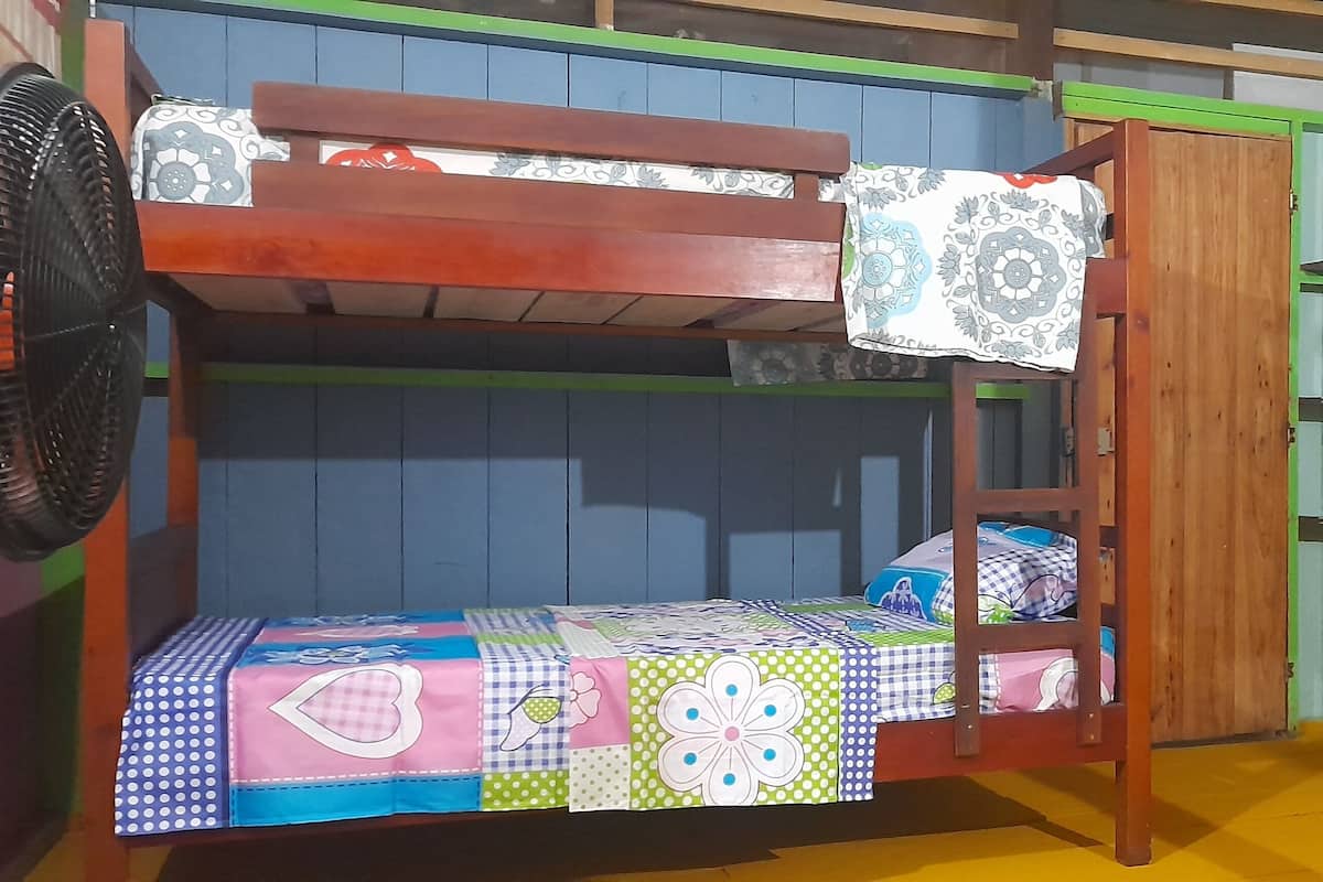 Un lit superposé avec une couverture aux couleurs vives, un ventilateur et une chaise en bois.