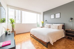 Una camera da letto moderna con un letto grande, pavimento in legno, una scrivania con un libro e piante in vaso.