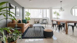 Een moderne woonkamer met een bank, salontafel, eettafel en stoelen. Er zijn grote ramen, een potplant en een hanglamp.