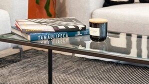 Un salon avec une table basse en verre, une plante en pot, un canapé agrémenté d’un coussin noir et blanc, et une pile de livres.