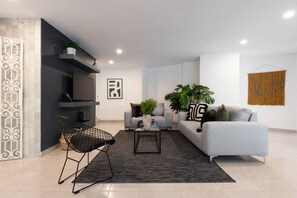 Un salon moderne avec un canapé gris, un fauteuil noir, une table basse et un téléviseur posé sur un meuble noir. On y trouve également deux plantes en pot et un tableau encadré accroché au mur.