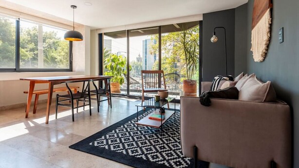 Un salon moderne comprenant un canapé, une table à manger, des chaises, un tapis à motifs et de grandes fenêtres donnant sur l’extérieur.