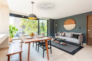 A modern living room with a dining area, a sofa, a round wall decoration, and a view of greenery outside.