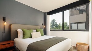 Une chambre moderne avec un grand lit, une table de chevet, une lampe fixée au mur et une vue sur les arbres par la fenêtre.