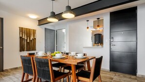 Un coin repas avec une table en bois dressée pour quatre personnes, entourée de chaises. Une porte noire se trouve à droite, et un luminaire fixé au mur est placé au-dessus de la table.