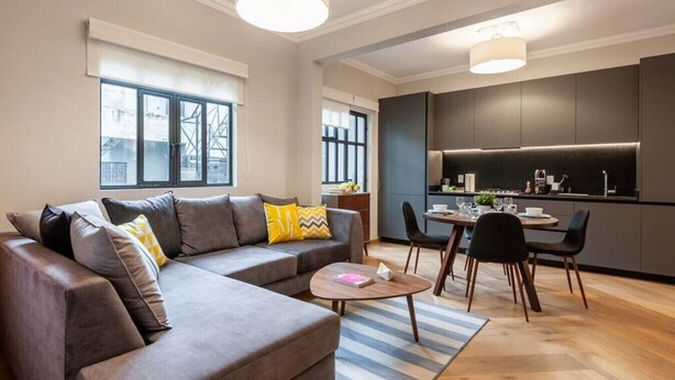 Un appartamento moderno con zona giorno che include un divano, un tavolino da caffè e un tavolo da pranzo con sedie. È presente un angolo cottura con mobili scuri e una finestra con vista sulla città.