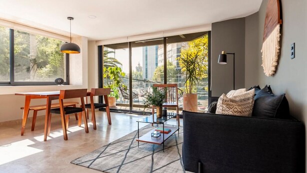 A modern living room with a black sofa, wooden chairs, a glass coffee table, and a large window offering a city view. - Wonderful 2bdr apt with beautiful natural light. (Ciudad de México)