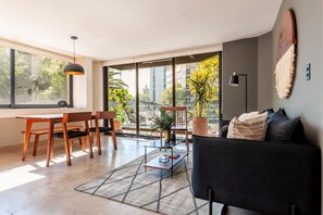 Un soggiorno moderno con un divano nero, sedie in legno, un tavolino da caffè in vetro e una grande finestra con vista sulla città.