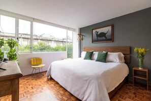 Ein Schlafzimmer mit einem großen Bett, Blick auf Grünfläche, ein gelber Sessel und ein Nachttisch aus Holz mit Blumenvase.