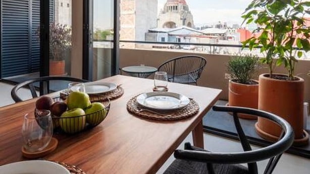 A dining area with a wooden table set for two, a modern pendant light, and a view of a cityscape with a prominent building.