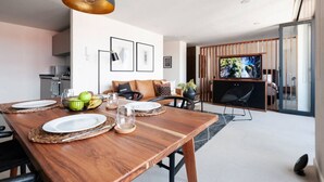 A modern dining area with a wooden table set for two, a black pendant light, and a television in the background.