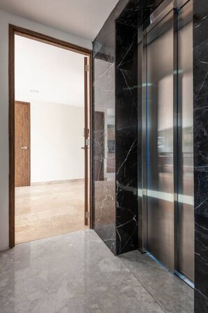 Un couloir avec une porte en bois donnant sur une pièce, un ascenseur en verre et un sol en marbre.