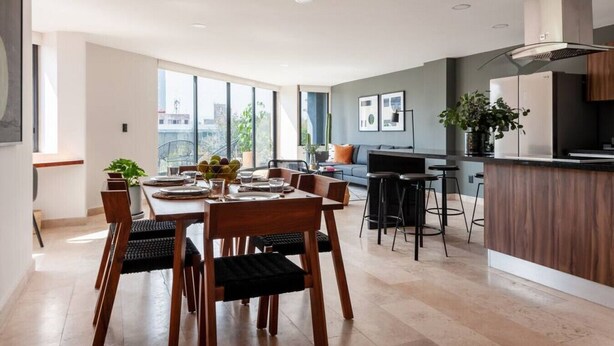 A modern kitchen with a central island, a dining area with wooden chairs and a table, and a living area with a sofa and a TV. - Lux large top floor Apt. at The Corner Condesa (Ciudad de México)