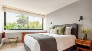 A modern bedroom with a large bed, a window with a view of trees, a bedside table with a lamp, and a small plant. - Lux large top floor Apt. at The Corner Condesa (Ciudad de México)