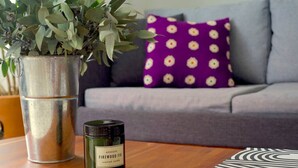 Ett vardagsrum med en grå soffa, en lila kudde med ett vitt blommönster, en grön växt i en metallkruka och ett litet grönt ljus märkt med texten "Firewood Fig" på ett träbord.
