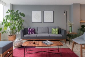 Un salon moderne avec un canapé gris, deux œuvres d’art encadrées, une table basse et une grande plante en pot.