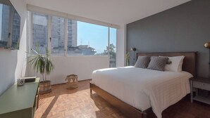 A modern bedroom with a large bed, a desk, a chair, a potted plant, and a view of buildings through a large window. - Darwin's Garden an amazing flat in Casa Quetzal (Ciudad de México)
