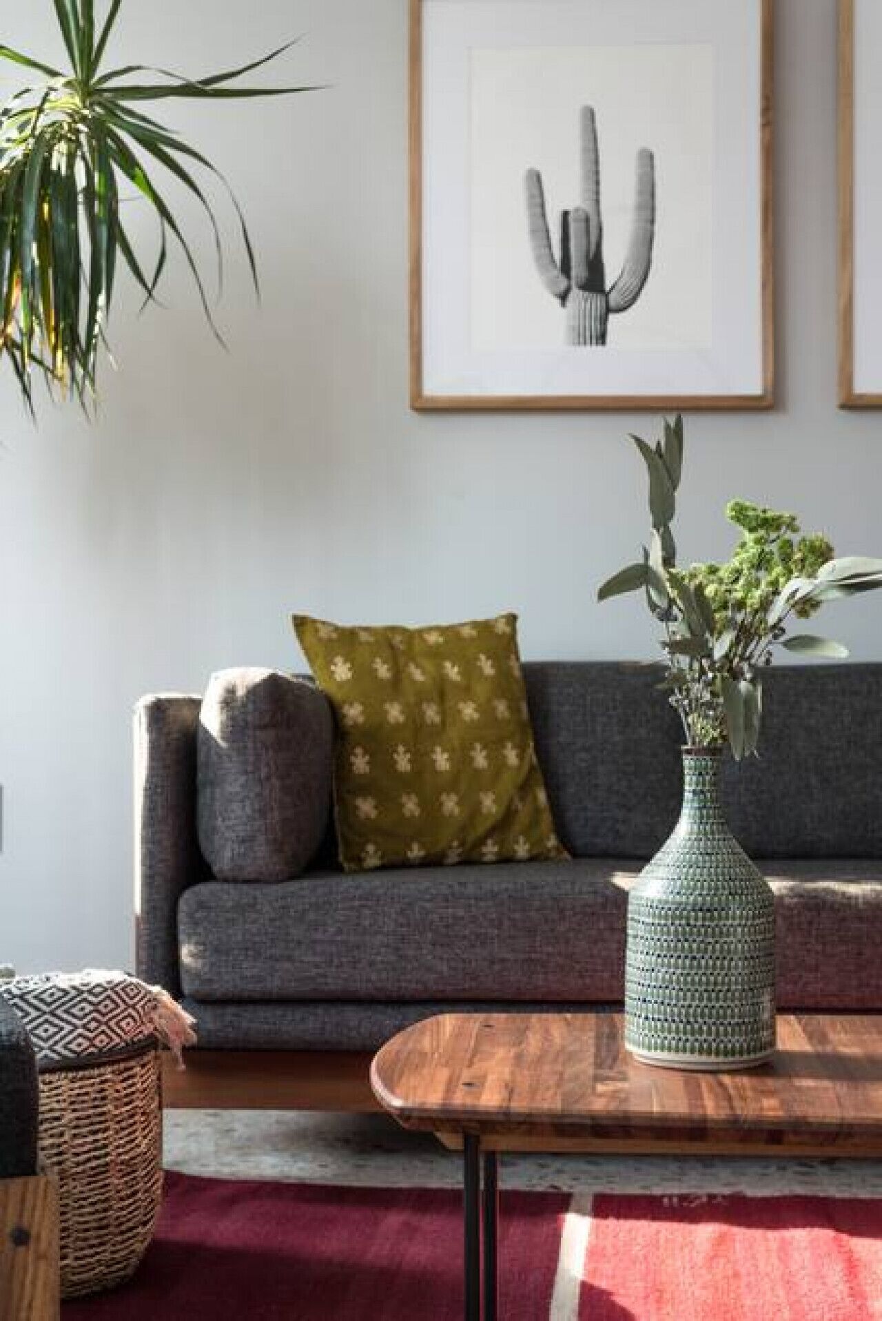 Un salon avec un canapé gris, un coussin vert à motifs, un vase vert contenant des plantes, une table basse en bois et une œuvre d’art encadrée accrochée au mur.