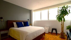 A bedroom with a bed, a nightstand, a potted plant, a window, and a radiator. - Bohemian Rooster, New! Deluxe Apt. in Casa Quetzal (Ciudad de México)