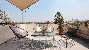 A rooftop terrace with two black wire-framed chairs, a glass table, and a patterned rug. A potted plant and a beige canopy provide shade. - Bohemian Rooster, New! Deluxe Apt. in Casa Quetzal (Ciudad de México)