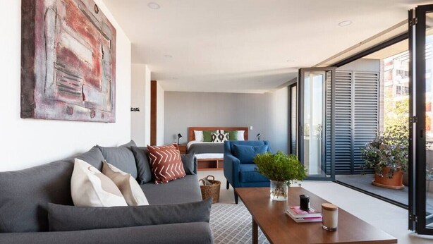Un salon moderne avec un canapé gris, un fauteuil bleu et une table basse en bois. Un tableau est accroché au mur et une plante en pot se trouve sur le balcon.