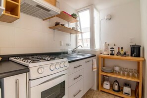 A compact kitchen with a gas stove, oven, and a sink. There are wooden shelves above the sink and a window providing natural light. - Mandarine Cardinal, Modern Apt. in Casa Quetzal (Ciudad de México)