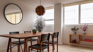 A dining area with a wooden table, six chairs, a hanging light fixture, a large mirror, and a window with a view.