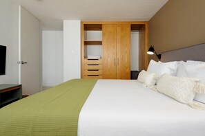 A bedroom with a bed, a green blanket, white pillows, a wooden wardrobe, and a TV on the left. - Mandarine Cardinal, Modern Apt. in Casa Quetzal (Ciudad de México)