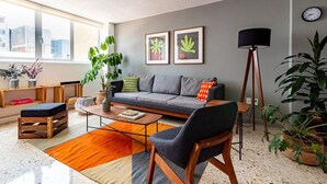Ein modernes Wohnzimmer mit grauem Sofa, Couchtisch und Stehlampe. An der Wand hängen zwei gerahmte Bilder, eine Zimmerpflanze und ein Bücherregal mit Büchern.