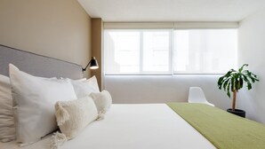 A hotel room with a bed, a bedside lamp, a window, and a potted plant.