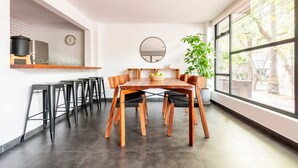 Un'area pranzo moderna con tavolo e sedie in legno, una grande finestra vista alberi, e un angolo cottura con una sveglia e una pentola nera sul piano di lavoro.