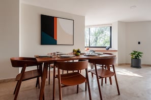 A dining area with a wooden table and chairs, a painting on the wall, a window with a view of trees, and a potted plant.