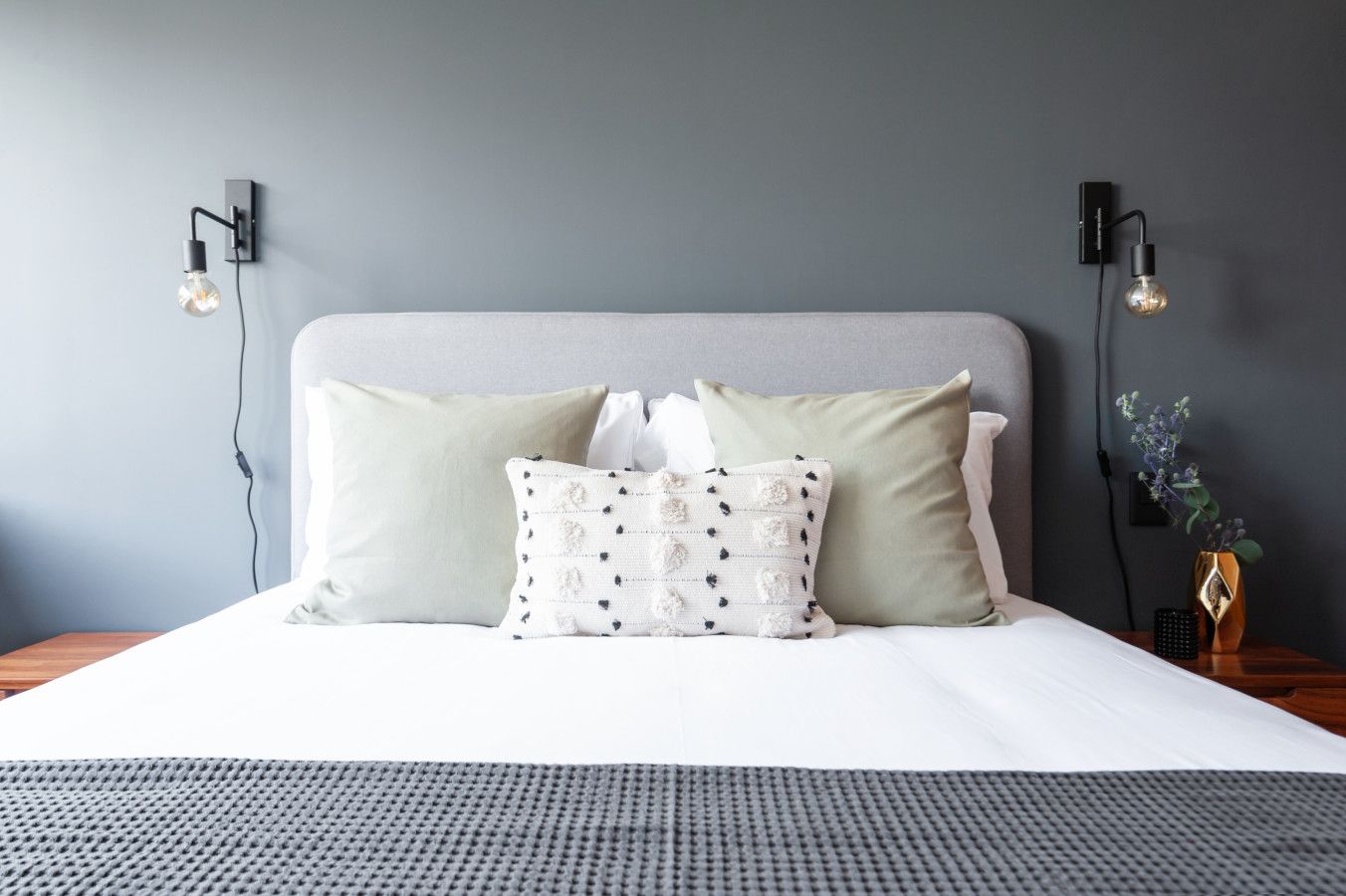 A bed with a grey headboard, white bedding, and decorative pillows. There are wall-mounted light fixtures on either side of the bed.