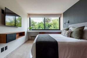 A modern bedroom with a large bed, a flat-screen TV mounted on the wall, and a window with a view of trees.