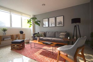 A modern living room with a grey sofa, a coffee table, a red patterned rug, a grey armchair, and a large plant.