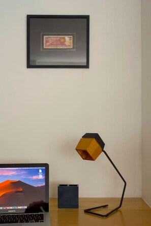 Ein Laptop auf einem Holztisch mit einem gerahmten Bild an der Wand und einer geometrischen Schreibtischlampe.