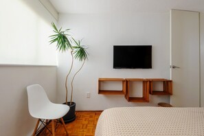 Ein Zimmer mit einem Bett, einem weißen Sessel, einem an der Wand befestigten Fernseher und zwei Topfpflanzen.