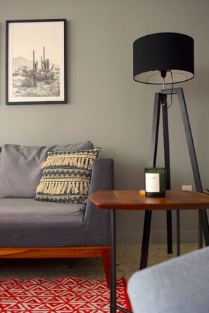 Una sala de estar con un sofá gris, cojines estampados, un tapete rojo y blanco, una lámpara de pie y una mesita con una vela.
