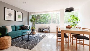 Un salon moderne avec un canapé de couleur turquoise, une table à manger avec des chaises, une table basse et une grande fenêtre donnant sur un paysage verdoyant.