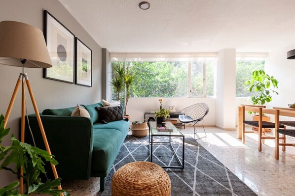 A modern living room with a green sofa, a coffee table, a chair, and a dining table. There are plants, a lamp, and framed pictures on the walls.