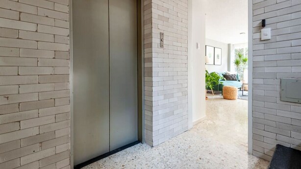 Un couloir avec une porte deascenseur fermée, des murs en briques blanches et un aperçu d’un salon avec un canapé et des plantes.