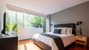 Une chambre avec un grand lit, une table de chevet, une commode, un téléviseur posé sur un meuble et une fenêtre donnant sur un paysage verdoyant.