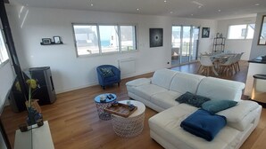Een ruime woonkamer met een witte bank, een salontafel en een eettafel met stoelen. Er zijn grote ramen die natuurlijk licht en uitzicht bieden.