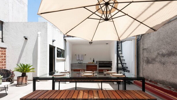 Un espacio exterior para comer con una mesa, bancos, un paraguas grande y una cocina al fondo.