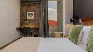 Une chambre d’hôtel moderne avec un grand lit, un bureau avec une chaise, une petite table ronde et une vue sur un immeuble rouge, visible à travers une fenêtre ornée de rideaux.