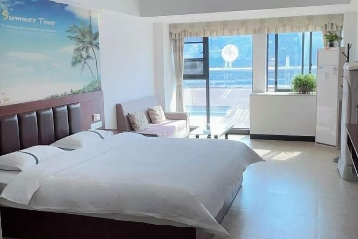A hotel room with a large bed, a small table, a sofa, a window with a view, and a refrigerator.